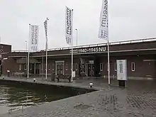  Eingangsbereich des Museum Rotterdam ’40 – ’50 NOW am Hafenbecken, mit mehreren Fahnen und einer Sitzbank vor dem Eingang