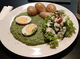 Grüne Soße mit einem halbier­ten hart­gekochten Ei und Pell­kartoffeln sowie einer Garnitur aus Blatt­salat, Tomaten­achteln und Dressing