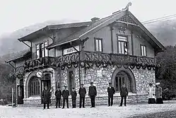 Schwarz-Weiß-Fotografie eines Bahnhofsgebäudes, davor mehrere in Uniform oder Arbeitskleidung gekleidete Personen