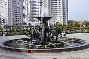 Ein runder Brunnen mit verschiedenen Figuren in der Mitte. Aus einer Art Schale die erhoben dargestellt ist, sprudelt Wasser.