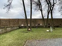 Im Vordergrund ist ein Weg und ein Wiesenabschnitt zu sehen. Mittig vom Bild befinden sich zwei Bäume, an deren Füßen die beige Gedenkkugel liegt. Den Hintergrund füllt die braune Lagermauer aus, an der verschiedene Gedenktafeln angebracht sind.
