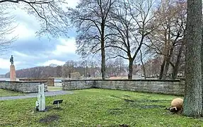 Links im Bild ist der See zu sehen, ebenfalls die auf einem sehr hohem Podest stehende Skulptur. Rechts im Bild befinden sich Bäume, an deren Fuß die Gedenkkugel liegt.