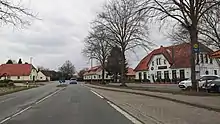Straßenszene, d. h. auf der Straße, ein Fahrzeug rund 50 Meter voraus. Links Häuser, rechts Häuser und eine Bushaltestelle sowie Speisegaststätte "Selinus"
