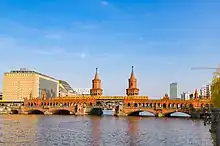 Frontale Farbfotografie von einer roten Backsteinbrücke mit einer gelben U-Bahn in der Mitte. Die Backsteinbrücke hat zwei große Türme und eine Unterführung für Autos und Fußgänger.