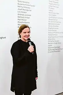 Brünette Frau mit Mikrofon vor einer Textwand mit Namen