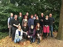 Gruppenbild mit 15 überwiegend fröhlich wirkenden Personen, im Hintergrund Wald