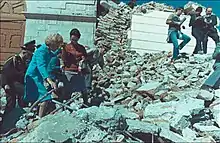 Pat Nixon, gestützt von einer Begleiterin, und Militärs klettern über Schutt durch Erdbebenschäden.