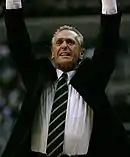 A man, wearing a jacket with a white shirt and tie, is raising his hands in the air during a basketball game.