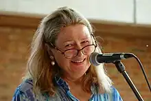 Porträt einer blonden Frau mit Brille, die den Kpopf leicht schräg hält, in ein Mikrofon spricht und das Publikum ansieht.