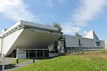 Frontale Farbfotografie eines Betongebäudes, aufgeteilt in zwei Komplexe. Links geht die vordere Mauer schräg nach hinten und hat oben eine schmale Fensterreihe sowie im Erdgeschoss einen Glaseingang. Rechts ist ein rechteckiger Gebäudeteil mit einem dreieckigen Aufbau, zwei unterschiedlich großen Fenstern und im Erdgeschoss eine Fensterreihe.