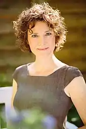 Ann Vielhaben hat kurze mittelbraune lockige Haare. Sie trägt ein kurzärmliges braunes Kleid vor einem sommerlichen Hintergrund und lächelt direkt in die Kamera