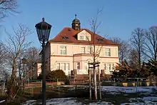In einer parkartigen Umgebung steht die Villa Storchennest, ein Gebäude mit zwei Stockwerken und einem Dachgeschoss unter dem rot eingedeckten Walmdach. Die sandfarbene Fassade ist mit ornamentalen Putzflächen nach Art des Jugendstils dekoriert. Ober- und Untergeschoss sind durch ein rötliches Gurtgesims deutlich voneinander getrennt, in der Mitte des Erdgeschosses springt ein Erker mit trapezförmigem Grundriss hervor. Eine Terrasse vor dem Erdgeschoss ist von roten Ziegelmauern umgeben und trägt ein Geländer aus weißen Holzlatten. In der Mitte des Daches, oberhalb des Erkers, gibt es eine größere Dachgaube mit Fenstern; darüber, auf dem Dachfirst, thront ein Türmchen. Es ist Winter, die Bäume sind kahl und die winterliche Sonne verleiht der Fassade einen warmen Gelbton.