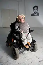 Zu sehen ist Raul Krauthausen in seinem Rollstuhl sitzend. Der Rollstuhl ist ein elektronischer und in dunklen Farben gehalten. Raul K.trägt beige Kleidung und eine helle Kappe.gtn