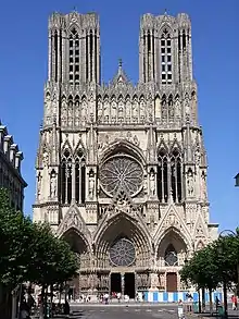 Frontale Farbfotografie einer gotischen Kirche mit zwei breiten, rechteckigen Turmspitzen. Viele kleine Türme, Säulen und Figuren verzieren das gesamte Gebäude. In der Mitte ist eine große Rosette, die von vier langen, schmalen Säulenfenstern begrenzt ist. Der Eingang besteht aus drei Spitzbögen, die mit Rosetten und vielen Figuren verziert sind.