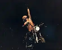 Halford mit freiem Oberkörper und zum Himmel gereckten Arm auf einem Motorrad