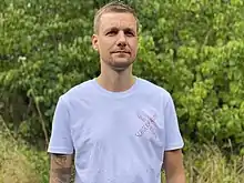 Tobias Schlegl steht im Grünen, bekleidet mit einem weißen T-Shirt, auf dem das Logo seines Romans „Schockraum“ abgedruckt ist.