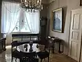 Farbige Eckansicht eines Innenraums mit einem runden Tisch, einem Kaffeeservice und drei Stühlen in der Mitte. An der linken Wand sind Fenster mit weißen Gardinen und einer verzierten Bank davor. Rechts vom Fenster befindet sich eine Standuhr. An der rechten Wand mit braun-weißem Muster hängt ein Porträt, unter dem ein Schreibtisch mit Büchern steht. Rechts davon eine weiße, verschlossene Tür. Am oberen Bildrand strahlt ein Kronleuchter.