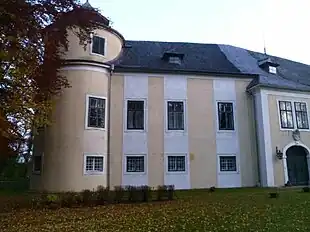 Links ein dreigeschoßiger, runder Eckturm, rechts ein Torbau mit Korbbogentor und gekuppeltem Fenster, dazwischen ein zweigeschoßiger Trakt mit drei Achsen und beige-weißem Anstrich
