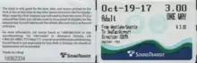 Busticket in Scheckkartenformat, Vorderseite und Rückseite nebeneinander abgebildet. Links, Rückseite, in kleiner Schrift Beförderungsbedingungen und Serviceinformationen. Rechts, Vorderseite, groß Datum Oct-19-17, Fahrpreis 3.00, Adult, One Way, kleiner From: Westlake/Seattle, To: SeaTac Airport, Directions:South. Unten Sound-Transit-Meereswellen-Farblogo.