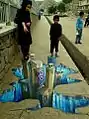 Auf dem Gehweg tut sich optisch ein Spalt auf. In Blautönen erscheint in einer Art Schlucht ein paar Hochhäuser. Auf dem Gehweg, an den "Rändern" dieser Schlucht stehen zwei Kinder.