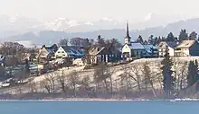 Tele-Aufnahme der Gemeinde Fehraltorf. Auf einem Hügel stehen einige Wohngebäude sowie die Kirche. Direkt unterhalb ist knapp der Pfäffikersee erkennbar.