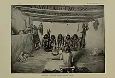 Das Bild zeigt eine gezeichnete indianische Zeremonie. Eine unbekleidete Person sitzt auf einem Hocker mit dem Rücken zum Zeichner. Vor ihr sitzen mehrere nackte Männer auf dem Boden, die Federn und kugelförmige Rasseln in den Händen halten. Vor ihnen stehen mehrere Holzfiguren auf dem Boden. Auf dem Boden im Zimmer befinden sich mehrere Kleidungsstücke, ein paar flache Schüsseln und ein kleiner weißer Teppich mit schwarzen Mustern am Rand. An der Wand hängen weitere Stofffetzen, zudem steht an einer Art Steinregal ein Webkamm, auf dem Regal liegen weitere Schüsseln.