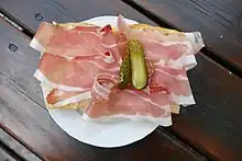 Fotografie eines Tellers, auf dem ein mit Speck und einer sauren Gurke belegtes Weißbrot liegt