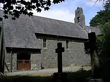 Teil einer steinernen Kirche mit hohem Schieferdach; links befindet sich eine leicht vorspringende Veranda mit hölzernen Türen, auf dem First befindet sich ein Glockenstuhl