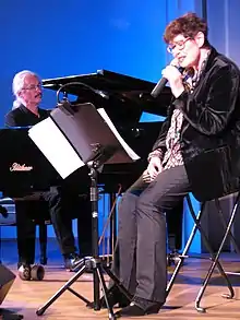 Uschi Brüning ist auf einem Klappstuhl sitzend vor einem Notenständer abgebildet. Sie trägt einen dunklen Blazer und eine graue Hose. In der linken Hand hält sie ein Mikrofon an ihren Mund.