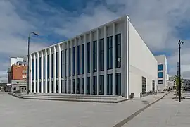 Foto eines weißen Gebäudes, auf der Vorderseite viele lange schmale Fenster, auf der Seite hingegen kaum Fenster