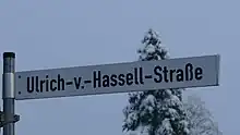 Foto eines weißen Straßenschildes vor grauem Winterhimmel mit der schwarzen Aufschrift Ulrich-v.-Hassel-Straße
