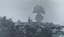 Schwarzweißfotografie von einer Stadt mit einer riesigen Rauchwolke. Einige Kirchtürme ragen hervor und vorne bedecken Bäume manche Häuser.