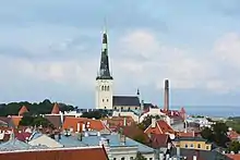Farbfotografie in der Obersicht von einer hellen Kirche, die von alten Häusern umgeben ist. Die dunkle Turmspitze ist teilweise oxidiert. Im rechten Hintergrund ist das Meer zu sehen.