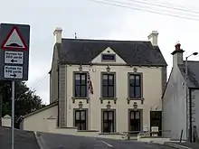 Ein zweistöckiges Gebäude mit hohen Fenstern, einer Plakette im Dachfirst und einer gehissten britischen Flagge davor.