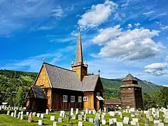 Foto einer Holzkirche mit Grabsteinen im Vordergrund und einer Erhebung dahinter