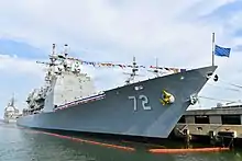 Über die Toppen geflaggter Kreuzer an Pier aus Richtung Wasser aufgenommen, Bug voraus. Kennnummer 72 zentral im Bild. Flagge Navy Jack, tiefblau mit den goldenen Sternen der Bundesstaaten, ist auf dem Vordersteven halb gehisst. Schiff ist von Ölbarrieren umgeben und liegt hoch im Wasser. Im Hintergrund der Achtersteven eines Schwesterschiffes und Masten anderer Schiffe.