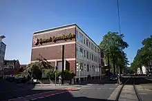 Ein Bild des Verwaltungsgebäudes der Privatbrauerei Herrenhausen. Dunkelbraune Backsteinfassade mit gelben Schriftzug "Brauerei Herrenhausen" in Frakturschrift und dem Logo der Brauerei, einem steigenden weißen Pferd auf rotem Grund.