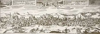 Kupferstich einer Stadt in der Frontalansicht mit den wichtigsten Gebäuden, die in zwei Legendenkästen am oberen Bildrand benannt sind. In der Mitte dieser Kästen steht in einem Banner „Vilna“. Hinter der Stadt ist eine hügelige Landschaft und im Vordergrund sind kleine Orte und Felder, auf denen Reiter und Menschen sind.