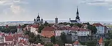 Farbige Panoramaaufnahme von einer Stadt mit einem Felsenberg in der Mitte, auf dem eine orthodoxe Kirche und ein Dom sind. In deren Mitte steht ein runder Burgturm. Viele alte Häuser und Türme mit braunen Dächern verzieren den Vordergrund, während hinten moderne Gebäude und Fabriken sind.