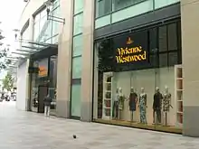 Seitliche Farbfotografie von einem Westwood-Geschäft in einem modernen Gebäude. Der gelbe Name Vivienne Westwood befindet sich mit gelbem Firmenlogo auf schwarzem Hintergrund und hängt über dem Eingang und im Schaufenster über sechs Schaufensterpuppen. Das gelbe Firmenlogo besteht aus einem Reichsapfel und dem ihn umkreisenden Planetenring.