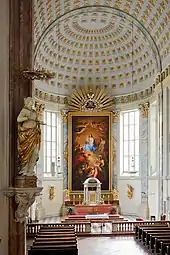 Frontale Farbfotografie eines Kirchenschiffs mit gold verzierter Bogendecke und einem hohen Gemälde hinter dem Altar. Das Zentrum des Gemäldes sind die Jungfrau Maria und das Christuskind, die von Menschen und Engeln umgeben sind. Über dem Gemälde sind Goldstrahlen und eine Krone zu sehen. Zwischen dem Gemälde und beiden Fenstern befinden sich helle Marmorsäulen. Auf dem Altar und an den Wänden sind goldene Figuren angebracht. Links vorne hängt an einem Pfeiler eine Marmorfigur mit goldenem Umhang und goldenem Heiligenschein.