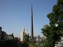 Blick über eine Eisenbahnstrecke auf einen teilweise metallverkleideter Gebäudekomplex in städtischer Umgebung. Das Foto wird in der Mitte von dem riesigen Backsteinschornstein dominiert. Links daneben ein deutlich niedrigerer, kastenförmiger, fensterloser Baublock mit heller Verkleidung und mehreren niedrigen, metallenen Schornsteinen über dem Dach.