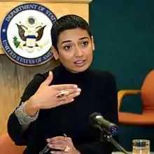 Salbi spricht in ein Mikrophon, sie hat kurze schwarze Haare, trägt einen schwarzen Pullover, sie macht erklärende Gesten mit beiden Händen, im Hintergrund ist das Emblem des US-Außenministeriums sichtbar