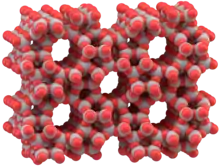 Eine Computergrafik aus grauen und roten Kugeln. Die Kugeln sind teilweise ineinander geschoben und bauen so eine räumliche Struktur auf. Graue Kugeln sind paarweise durch jeweils eine rote Kugel verbunden. In der Struktur sind fünf zylindrische Hohlräume, um die herum sich das Netzwerk der Kugeln legt. Die Hohlräume sind untereinander symmetrisch angeordnet und ihr Durchmesser ist in etwa genau so breit wie der Abstand zwischen zwei Hohlräumen.