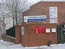 Das Ruderhaus ist mit Ziegelsteinen erichtet. Auf einer Tafel steht Bundesleistungszentrum und Landesleistungszentrum Rudern. Darunter ist die Tafel der Rudervereinigung mit den beiden gekreuzten Fahnen erkennbar. Das Gelände ist von einer Ziegelmauer umgeben, auf dem Boden liegt Schnee.