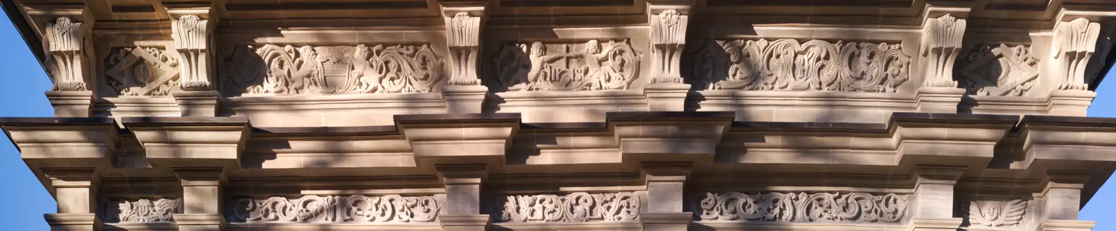 Rankenfriese im Gebälk der Südseite mit folgenden Motiven: unten gegen die Aussenkante: je eine Putte. Oben (von links nach rechts): Raute mit Mond, von zwei Löwen gehaltenes Freiburger Staatswappen, von zwei Engeln präsentiertes Jesuitenwappen, nicht identifiziertes Wappen (eventuell Zimmermann) und Raute mit der strahlenden Sonne.