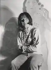 Maria Lassnig auf einem selbst fotografierten Schwarzweißbild von 1974, im Hintergrund das Gemälde Doppelselbstportrait mit Kamera